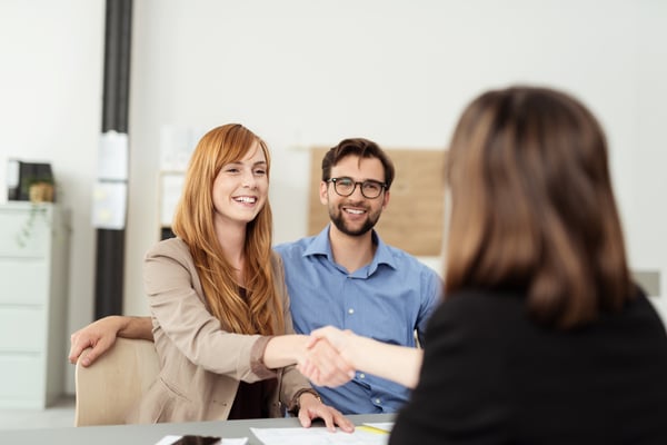 Happy young couple meeting with a broker in her office leaning over the desk to shake hands, view from behind the female agent